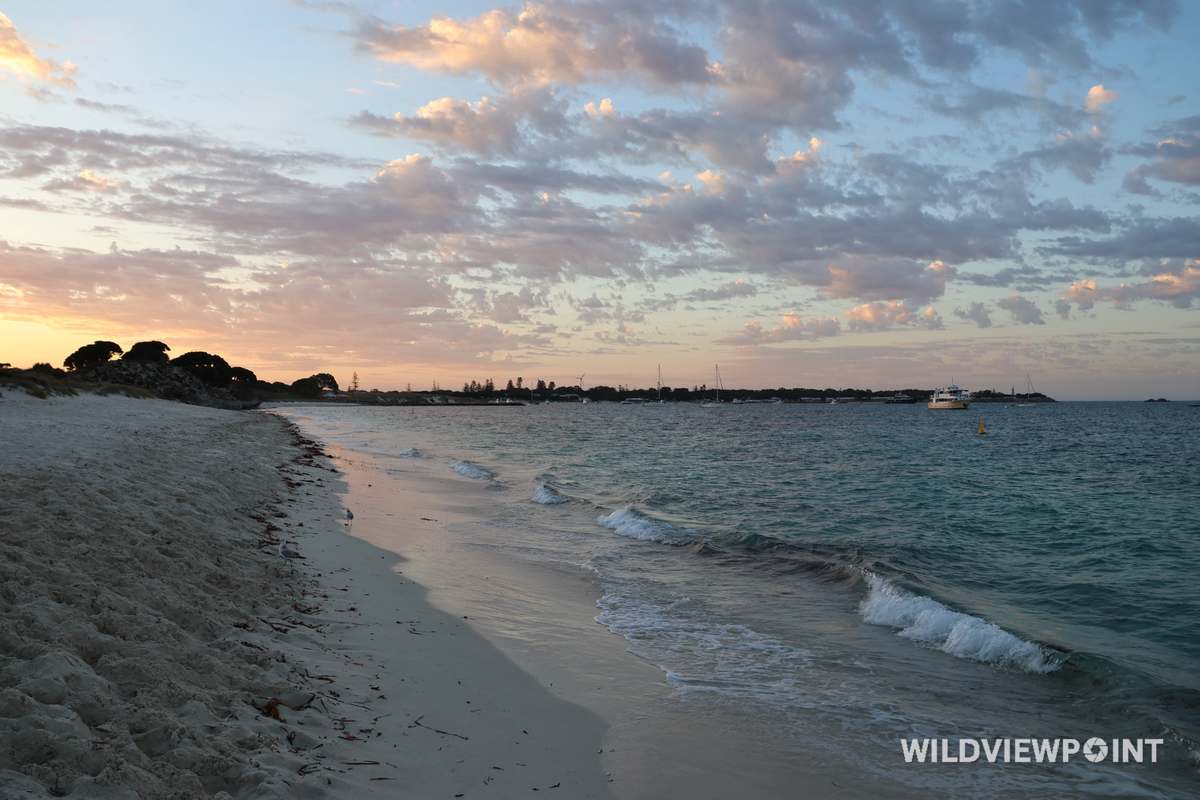 A white sand beach with small waves under pink-lit clouds, with moored boats and a ferry in the distance.