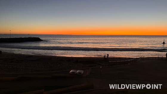 A bright orange band along the horizon above a rock groyne, with people fishing in silhouette and small waves breaking on a dark beach.