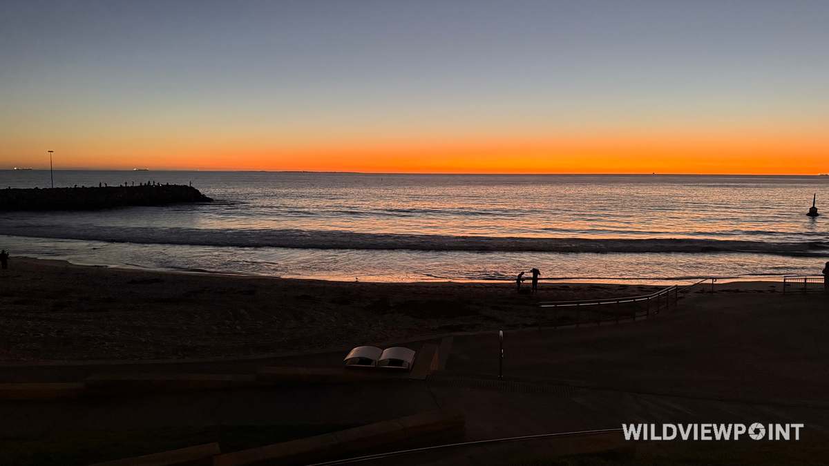 A bright orange band along the horizon above a rock groyne, with people fishing in silhouette and small waves breaking on a dark beach.