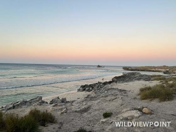 A sandy beach with weathered rocks and dune grass looking out over breaking waves, under a pale peach and blue sky.