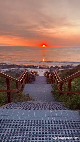 A grated stairway with timber handrails runs down toward the sea as a red sun sets on the horizon, its reflection in the water.