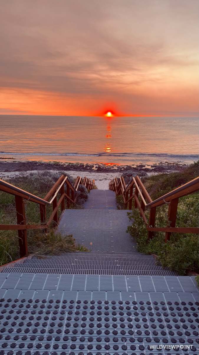 A grated stairway with timber handrails runs down toward the sea as a red sun sets on the horizon, its reflection in the water.