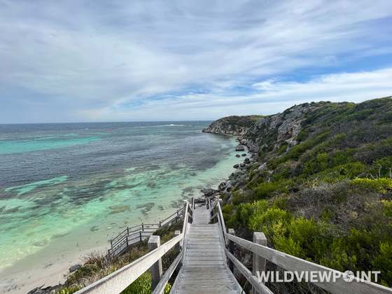 A weathered timber staircase descends a green coastal hillside to a bay of turquoise water over reef.