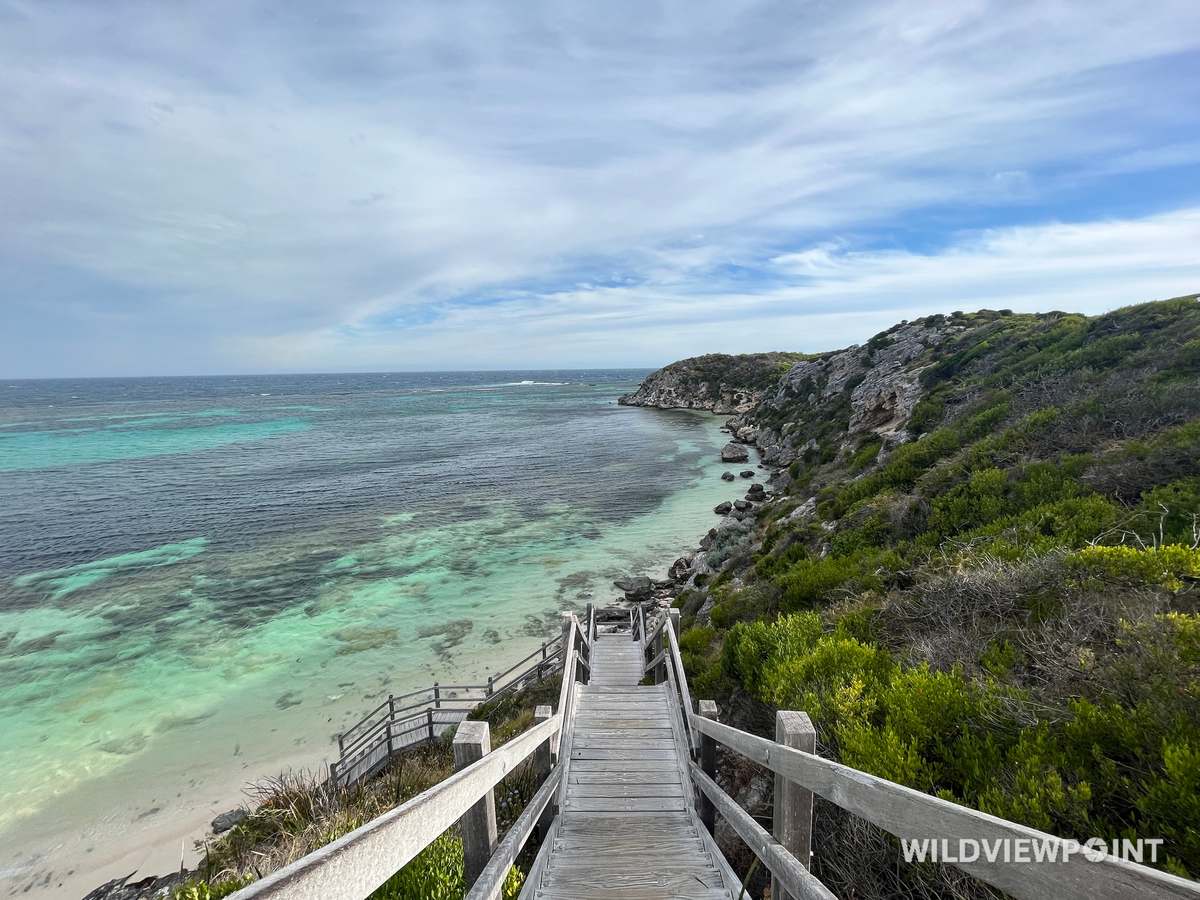 A weathered timber staircase descends a green coastal hillside to a bay of turquoise water over reef.