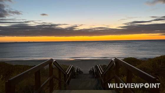 A timber stairway leads down between handrails to a calm beach, under a blue dusk sky with an orange band at the horizon and ships on the water.
