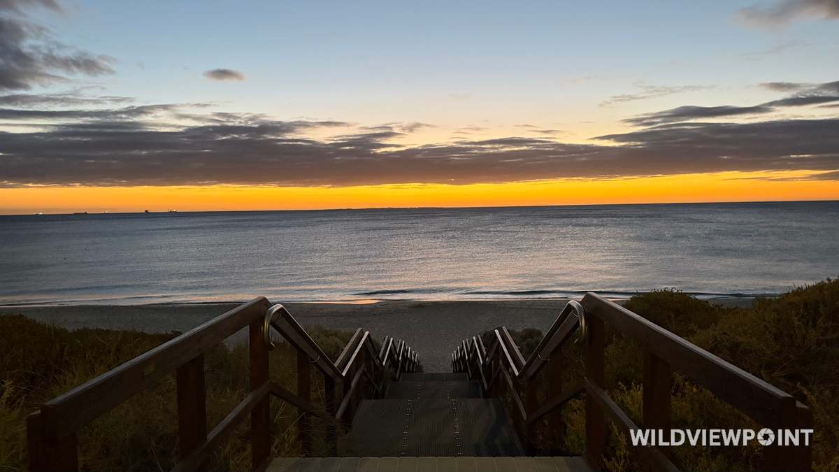 A timber stairway leads down between handrails to a calm beach, under a blue dusk sky with an orange band at the horizon and ships on the water.