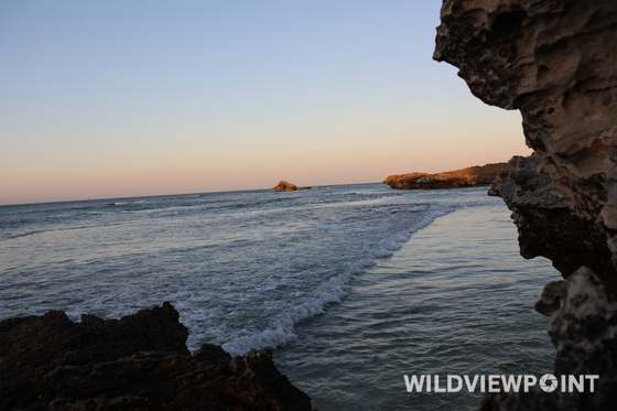 Surf running between weathered limestone rocks in warm evening light, a rock stack in the distance.