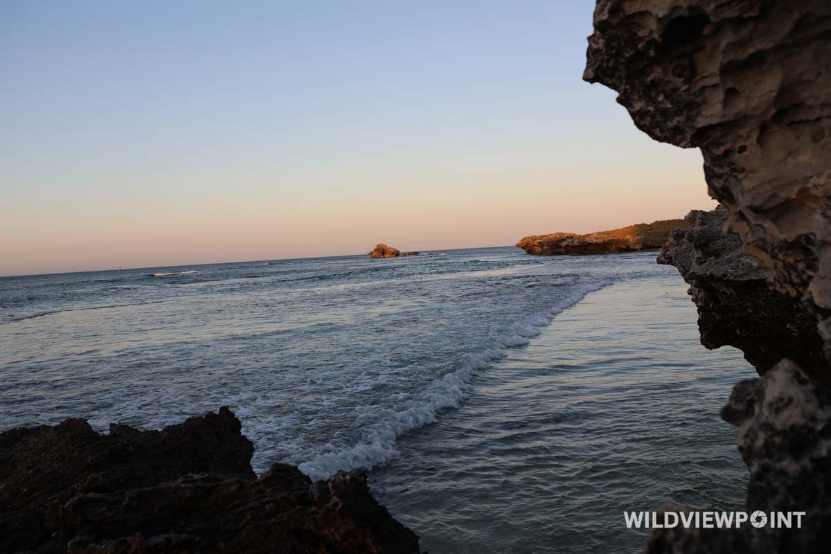 Surf running between weathered limestone rocks in warm evening light, a rock stack in the distance.